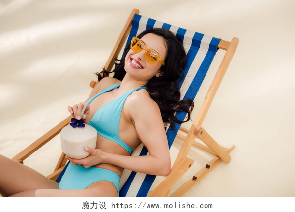 一个女人躺在沙滩躺椅上手里拿着椰子汁美丽的快乐女孩在比基尼和太阳镜与椰子鸡尾酒躺在甲板上的椅子上海滩